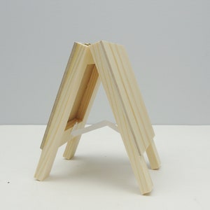 May include: A small wooden easel with a natural finish. The easel is made of two pieces of wood that are joined together at the top. The easel is designed to hold a small canvas or picture.