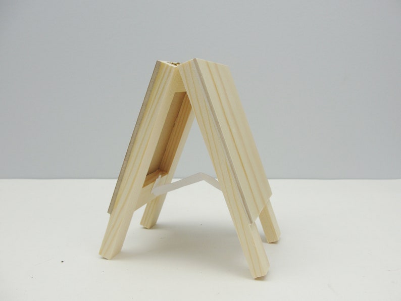 May include: A small wooden easel with a natural finish. The easel is made of two pieces of wood that are joined together at the top. The easel is designed to hold a small canvas or picture.