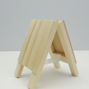 May include: A wooden easel with a natural finish. The easel is made of two pieces of wood that are joined together at a right angle. The easel is standing on a white surface.