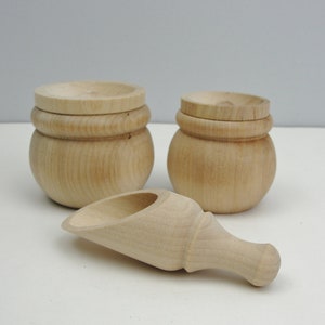 May include: Two wooden containers with lids and a wooden scoop. The containers are round and have a smooth, natural finish. The scoop is curved and has a handle.