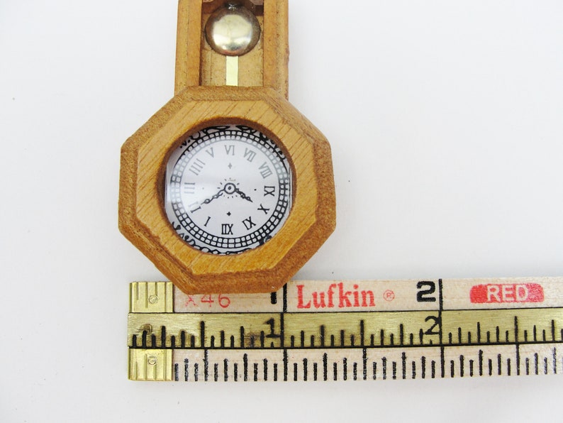 May include: A small wooden clock with a white face and black Roman numeral markings. The clock is about 2 inches in diameter and is being measured with a ruler.