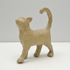 May include: A tan-colored paper mache cat figurine. The cat is standing with its tail curved upwards. The cat's ears are perked up, and it appears to be looking upwards. The figurine is made of a textured, natural-looking material.