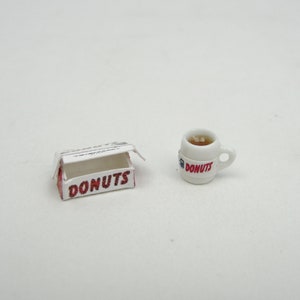 May include: A miniature white box with the word DONUTS printed in red on the front, accompanied by a small white mug featuring a handle and the word DONUTS in red text, containing a light brown liquid with a small white object inside.