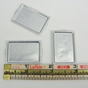 May include: Three silver baking sheets, measuring approximately 1 inch by 2 inches, are displayed on a white surface with a ruler for scale.