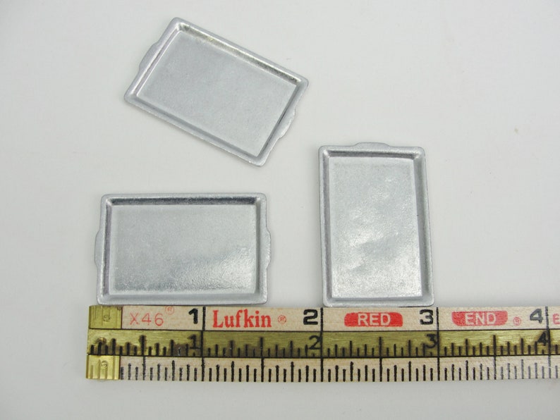 May include: Three silver baking sheets, measuring approximately 1 inch by 2 inches, are displayed on a white surface with a ruler for scale.