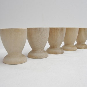 May include: Five wooden egg cups in a row on a white surface. The cups are all the same size and shape, and they are made of light-colored wood.