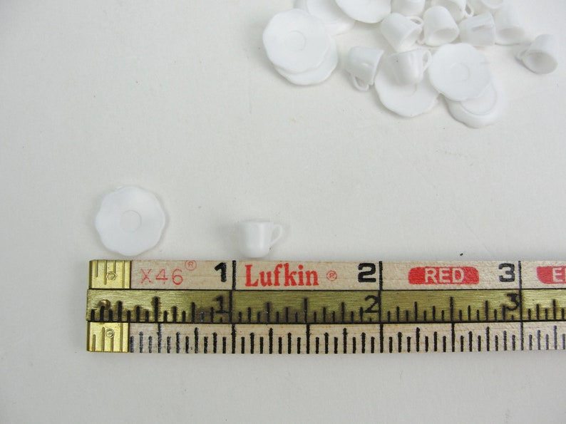 May include: A white ceramic teacup and saucer set, measuring approximately 1 inch in diameter, next to a wooden ruler marked with the brand "Lufkin" and the numbers 1, 2, and 3.
