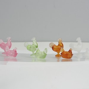May include: A collection of colorful glass rooster and hen figurines in pink, green, orange, and white. The rooster figurines stand upright, while the hens are in a sitting position. The figurines are displayed on a white surface.