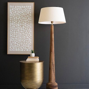 May include: A gold metal side table with a round top and a wide base. A wooden floor lamp with a white lampshade sits next to the table. A framed abstract art print with white shapes on a beige background hangs on the wall behind the table and lamp.