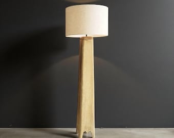May include: A tall, wooden floor lamp with a rectangular base and a beige lampshade. The lamp has a natural wood finish and stands on a concrete floor against a dark gray wall. The lampshade is cylindrical.