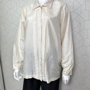 Op de afbeelding: Een crèmekleurig shirt met lange mouwen, een kraag en een knoopsluiting. Het shirt heeft een getextureerde, licht gekreukelde uitstraling en wordt gecombineerd met een zwarte broek. De mannequin wordt gepresenteerd tegen een geometrisch patroon.