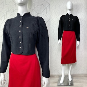 Puede incluir: Chaqueta negra de lana con botones plateados y costuras decorativas, combinada con una falda roja. La chaqueta tiene cuello alto y mangas largas. El conjunto se exhibe en un maniquí.