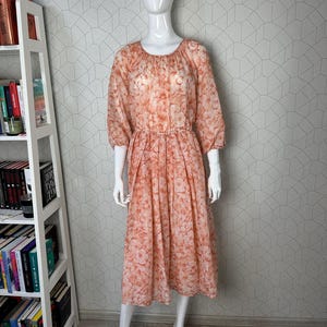 May include: A peach-coloured midi dress with a floral print. The dress has a round neckline, 3/4 sleeves, and a gathered waist.
