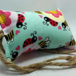 May include: A small, light blue pillow with a pattern of ladybugs, bees, and flowers. The pillow has a brown twine tie.