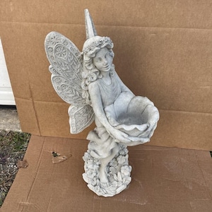 May include: A gray concrete fairy statue with detailed wings and a floral crown. The fairy holds a large, open bowl, likely for a bird bath or decorative display. The statue is on a base with floral accents.