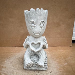 May include: A gray concrete statue of a character holding a heart shape in its hands. The character has a large head with a wide smile and large eyes.