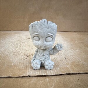 May include: A gray concrete sculpture of a character with large eyes and a small smile, raising one hand in a wave. The figure has a textured, tree-like appearance, with detailed facial features and a small body. The sculpture is a decorative item.