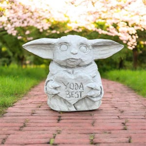 May include: A light gray statue of a character holding a heart-shaped sign that reads "YODA BEST." The statue has large ears and is set on a brick pathway with a blurred green and pink background.