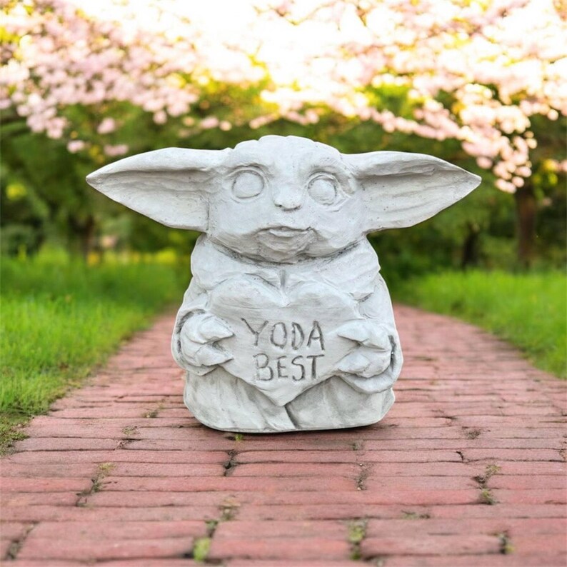 May include: A light gray statue of a character holding a heart-shaped sign that reads "YODA BEST." The statue has large ears and is set on a brick pathway with a blurred green and pink background.