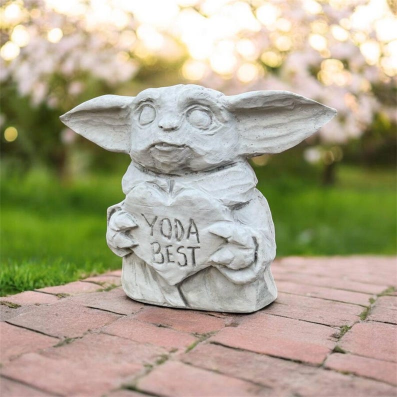 May include: A gray concrete statue of a character holding a heart-shaped sign that reads "YODA BEST". The statue has large ears and is set on a brick patio with a blurred green and white background.