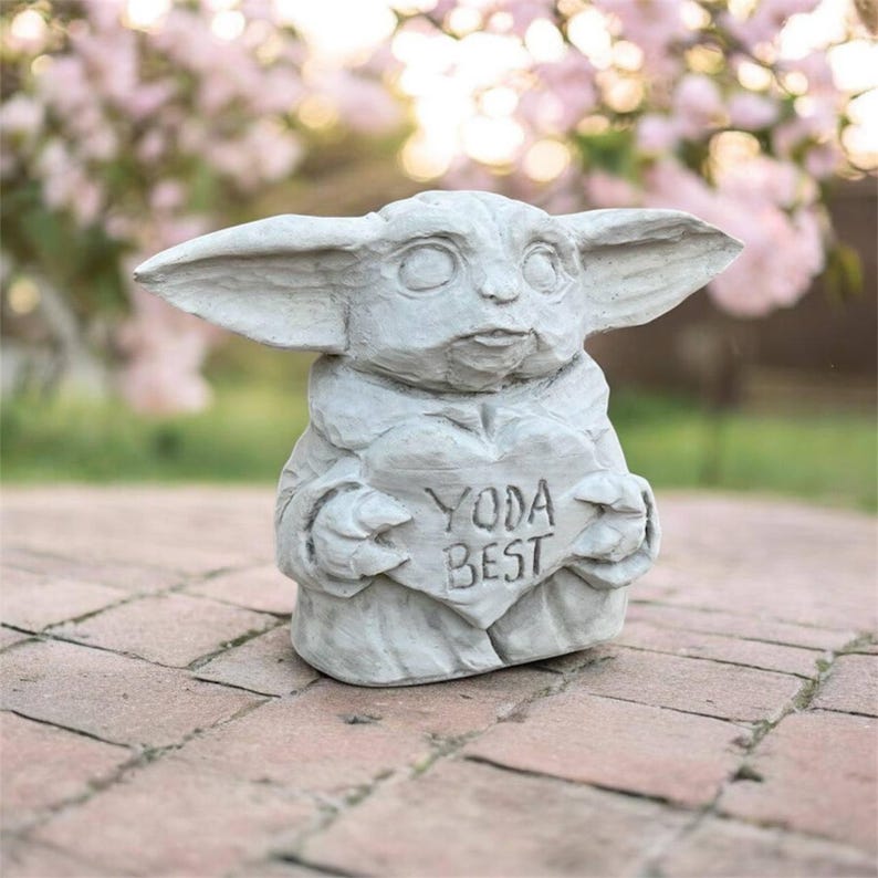 May include: A gray statue of a character with large ears, holding a heart-shaped sign that reads "YODA BEST". The statue is made of a stone-like material and is set on a brick surface, with a blurred floral background.