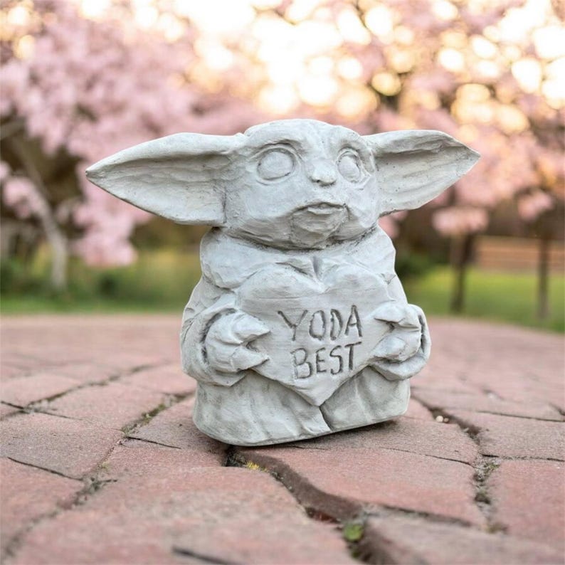 May include: A gray concrete statue of a character with large ears, holding a heart-shaped sign that reads "YODA BEST". The statue is set on a brick surface with a blurred background of pink flowering trees.