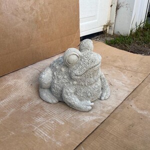 May include: A gray concrete frog statue with a textured surface and large eyes. The frog is sitting on a brown cardboard surface.
