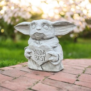 May include: A gray concrete statue of a character holding a heart-shaped sign that reads "YODA BEST". The statue has large ears and is set on a brick patio with a blurred green and white background.