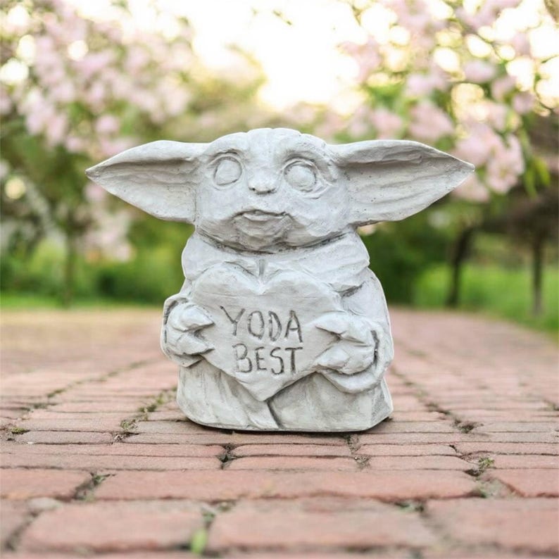May include: A gray statue of a character holding a heart-shaped sign that reads "YODA BEST." The statue has large ears and is set on a brick pathway with a blurred background of pink flowers.