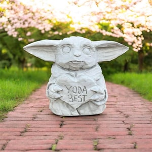 May include: A light gray statue of a character holding a heart-shaped sign that reads "YODA BEST." The statue has large ears and is set on a brick pathway with a blurred green and pink background.