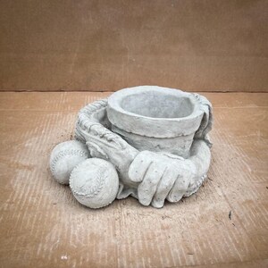 May include: A concrete planter shaped like a baseball glove holding two baseballs. The planter has a round opening for planting.