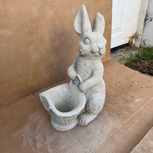 May include: A gray concrete rabbit statue holding a basket planter. The rabbit has a sweet expression and is standing on a brown surface.