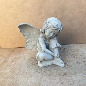 May include: A gray concrete fairy statue with detailed wings, a floral headpiece, and a seated pose. The fairy is looking down, with her arms wrapped around her legs. The statue is designed for outdoor garden decor.