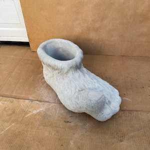 May include: A gray concrete planter shaped like a furry foot. The planter has a round opening at the top for planting.