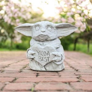 May include: A gray statue of a character holding a heart-shaped sign that reads "YODA BEST." The statue has large ears and is set on a brick pathway with a blurred background of pink flowers.