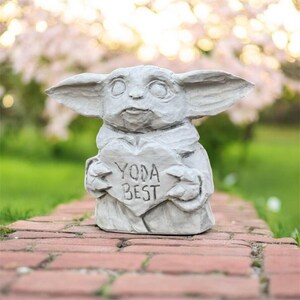 May include: A gray concrete statue of a character with large ears, holding a heart-shaped sign that reads "YODA BEST". The statue is set on a brick surface with a blurred background of green grass and pink flowers.
