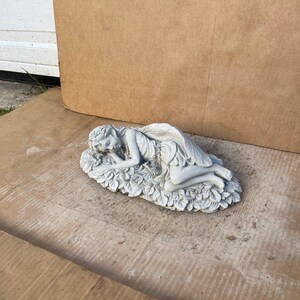 May include: A light gray fairy statue, sculpted in a reclining pose on a bed of leaves. The fairy has detailed wings and a flowing dress. The statue is made of a durable material, suitable for outdoor display.