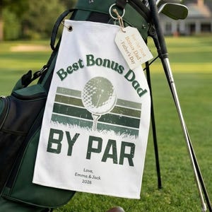 以下が含まれることがあります： 緑色のゴルフバッグとクラブ、白いタオルには「Best Bonus Dad BY PAR」の文字、芝生にはゴルフアクセサリーがあります。タオルにはゴルフボールのグラフィックと「Love, Emma & Jack 2026」の文字があります。