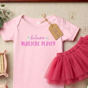 May include: A light pink short-sleeved baby bodysuit with the text "future MAHJONG PLAYER" in purple and pink, featuring two small mahjong tile graphics. A brown cardboard tag labeled "CURATED with CARE" is attached to the neckline.