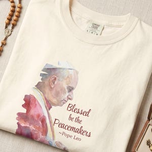 May include: Cream-coloured t-shirt with a watercolour image of Pope Leo and the text "Blessed be the Peacemakers." A wooden rosary and a gold-coloured dove pendant in a wooden box are also visible.