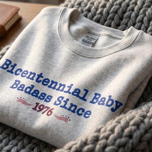 May include: A light grey sweatshirt featuring the text "Bicentennial Baby Badass Since 1976" in blue and red. The sweatshirt is made of a soft material and is folded on a grey knitted blanket.