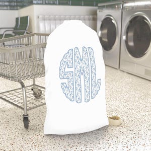 May include: White laundry bag with a light blue floral monogram of the letters SML. The bag is made of a soft material and is standing on a speckled floor. A metal laundry cart is in the background.
