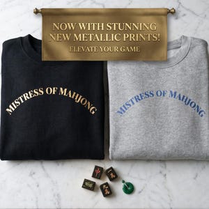 May include: A black sweatshirt with gold text reading MISTRESS OF MAHJONG and a gray sweatshirt with blue text reading MISTRESS OF MAHJONG. Above them, a banner reads NOW WITH STUNNING NEW METALLIC PRINTS! ELEVATE YOUR GAME. Four mahjong tiles and a green jade ring are below.