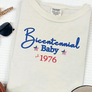 Puede incluir: Camiseta color crema con el texto "Bicentennial Baby est 1976" en azul y rojo. El dise&ntilde;o incluye estrellas y rayas, celebrando el a&ntilde;o 1976.