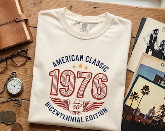 Puede incluir: Camiseta color crema con un diseño vintage. La camiseta presenta el texto "AMERICAN CLASSIC 1976 BICENTENNIAL EDITION" en azul marino y rojo. Una pequeña etiqueta dice "A True Classic - Happy 50th!"