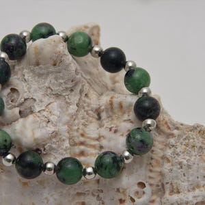 Könnte beinhalten: Ein grünes und schwarzes Edelstein-Armband mit silbernen Perlen. Das Armband liegt auf einer weißen Muschel.