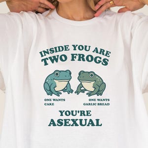 Puede incluir: Camiseta blanca con el texto "INSIDE YOU ARE TWO FROGS" sobre dos ranas de dibujos animados. Debajo de las ranas, el texto dice "ONE WANTS CAKE" y "ONE WANTS GARLIC BREAD". La frase "YOU'RE ASEXUAL" está impresa debajo.