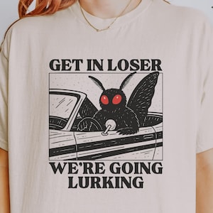 Pode incluir: Camiseta bege com um gráfico em preto e branco de uma mariposa dirigindo um carro. O texto "GET IN LOSER" está acima do carro e "WE'RE GOING LURKING" abaixo. A camiseta tem gola redonda e mangas curtas.