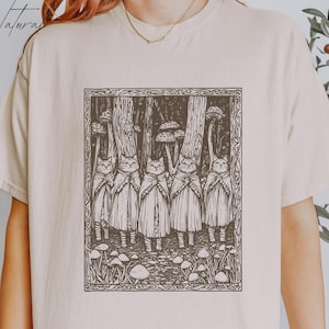 Puede incluir: Una camiseta beige con una ilustración en blanco y negro de un grupo de gatos con capas de pie en un bosque con setas. La ilustración está enmarcada con un borde negro.