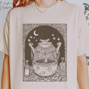 Puede incluir: Una camiseta blanca con un gráfico marrón de una rana con una túnica y sosteniendo un caldero, con estrellas y una luna creciente en el fondo.